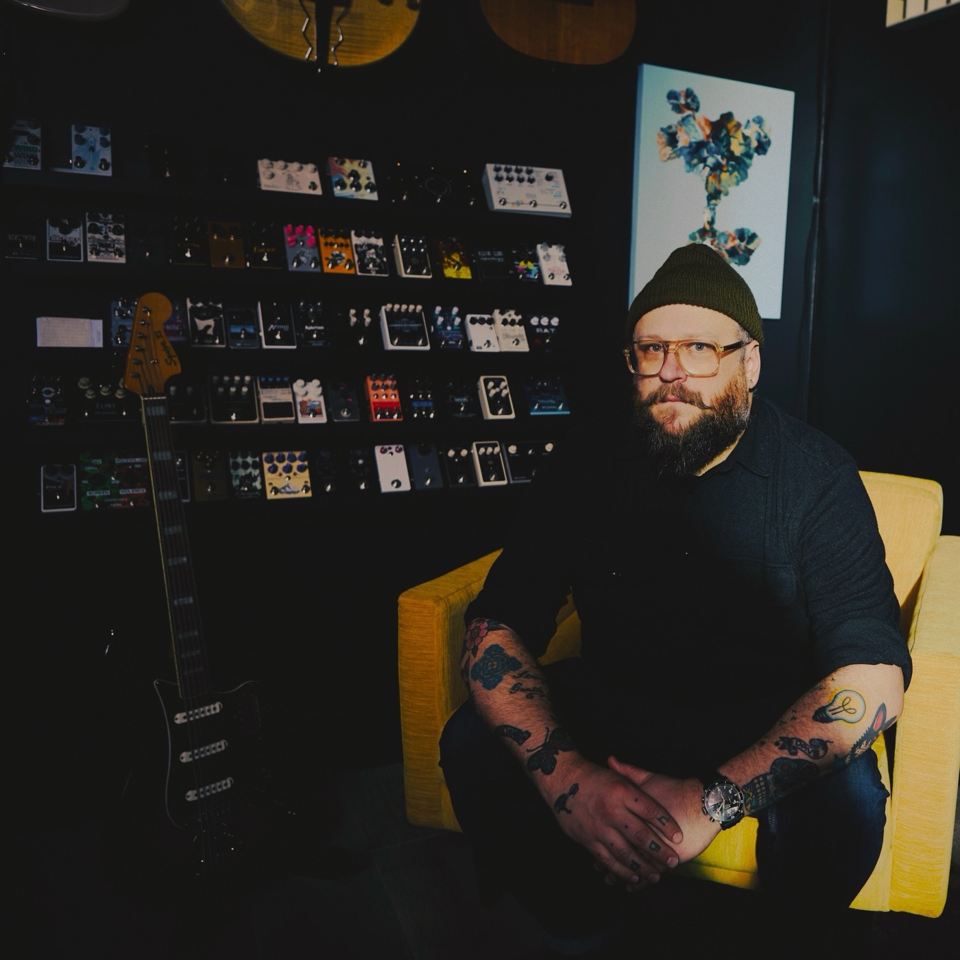 Daniel G. Harmann seated in a yellow armchair in front of a wall of guitar effects pedals, an electric guitar leaning beside him.